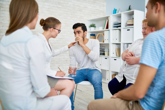 Portrait of female psychiatrist comforting crying young man in group therapy session with other patients sitting in circle around them