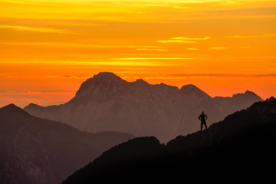 Happy successful winning man reaching mountain summit. Spectacular layered mountain ranges silhouettes with orange sunset sky.