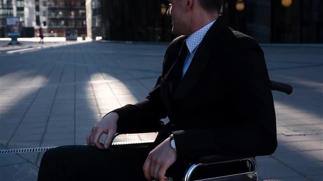 Slow motion shot of a businessman with in wheelchair walking and turning around abd look at camera on the business district square. Happy smiling cripple man. Portrait.