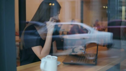 View from the street of an attractive young woman talking on the phone in a cafe and looking at the screen of her laptop during her coffee break by the window in the cafe