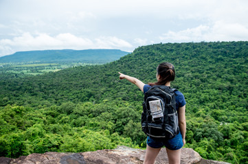 Young woman with backpack map behind bag standing on cliff's edge pointing to sky