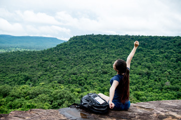 Young woman with backpack sitting on cliff's edge looking to sky with raised hands and enjoy morning