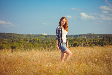 Naklejka na ściany i meble Red-haired attractive American style girl, checkered shirt with a white top and shorts standing in a meadow at sunset enjoying the rays of the sun