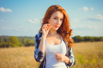 Red-haired attractive American style girl, checkered shirt with a white top and shorts standing in a meadow at sunset enjoying the rays of the sun