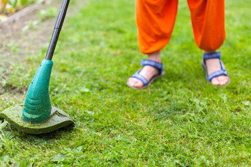 Gasoline lawn trimmer mows juicy green grass on a lawn on a sunny summer day. Close-up selective focus image. Garden equipment. Young woman mowing the grass with a trimmer.