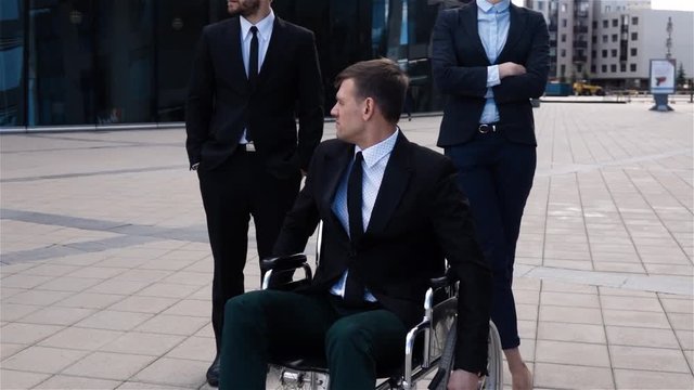 Epicly of disease disability paralysis handicap health concept. Handicapped male with disabled legs sitting on wheelchair. His colleagues standing back and turn around head to camera.