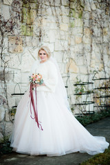 Obraz premium Magnificent bride in luxury dress and white coat stands before stone wall