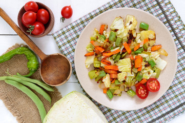 Vegetable stew - a mixture of baked cabbage, green beans, onions, carrots, cherry tomatoes, sweet pepper on a plate on a white wooden background. Vegan cuisine. Proper nutrition.