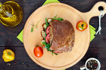 Big sandwich with pieces of meat, arugula, tomato, cereal ciabatta on a cutting board on a dark wooden background. Top view.