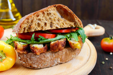 Big sandwich with pieces of meat, arugula, tomato, cereal ciabatta on a cutting board on a dark wooden background.