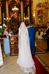 Naklejka premium Look from behind at groom in blue suit and bride in long dress wtih long veil standing in church