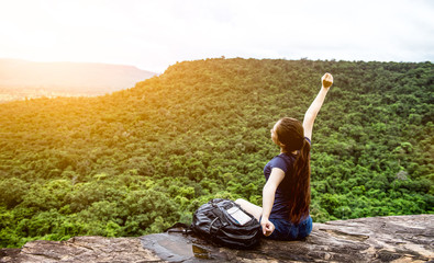 Naklejka na ściany i meble Young woman with backpack sitting on cliff's edge looking to sky with raised hands and enjoy morning sunrise