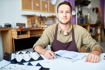 Portrait of smiling young man sitting at table in jeweler shop looking at camera with documentation and ring  set in front of him