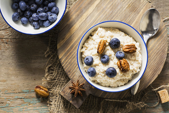 Healthy breakfast: milk porridge from oat bran on skimmed milk with honey, juicy blueberries, pecans on a simple wooden background with a spoon and a jar of honey. Top View.