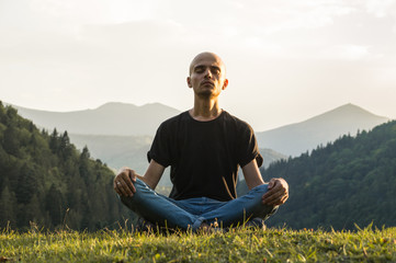 Young man does yoga in mountains at sunset. Skinny male person sits in lotus position top of the...