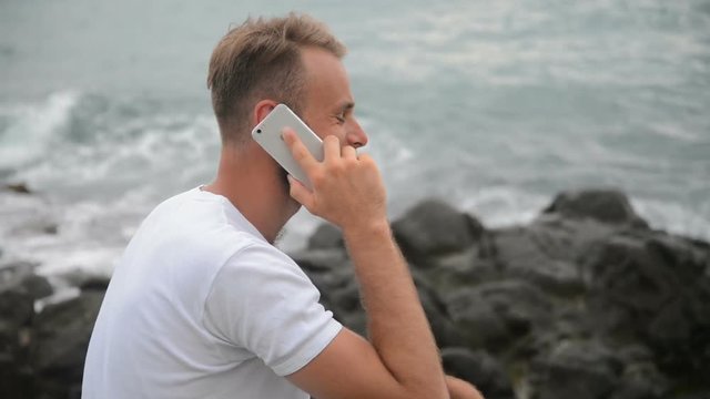 Young man talking on cellphone standing on beach near sea