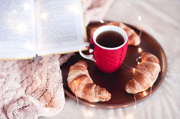 Tasty breakfast with fresh croissants and black tea staying on wooden tray with open book in bed closeup. Good morning.