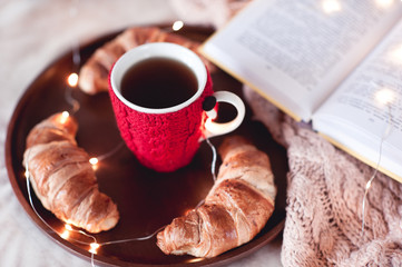 Knitted cup of black tea with homemade croissants and open book staying on wooden tray in bed closeup. Winter season. Good morning.