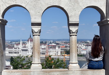 A view of the Hungarian Parliament at Danube river embankment in Budapest through the arches of Fishermen Bastion of Buda Castle. Tourist girl sitting in the window.