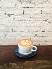 Cup of hot milk latte coffee with rosetta latte art on wooden table background and white brick wall. Concept of morning drink freshness