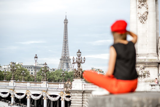 Young woman in red hat enjoying great view on Eiffel tower sitting on the bridge in Paris. Image focused on the background