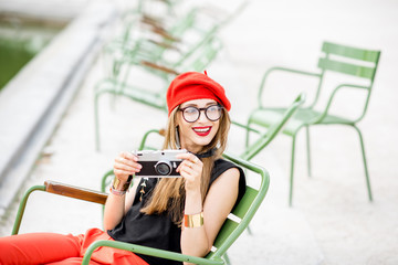 Fototapeta na wymiar Young stylish woman tourist in red cap and pants sitting with photocamera on the famous green chairs in Tuileries garden in Paris