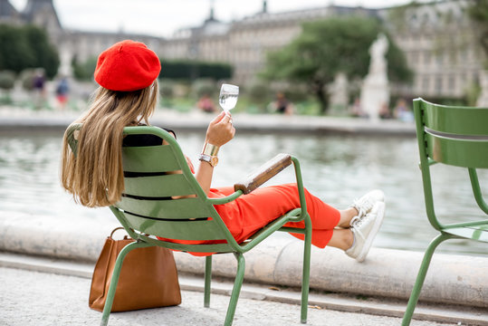 Young stylish woman in red cap and pants sitting with glass of wine on the famous green chairs in Tuileries garden in Paris
