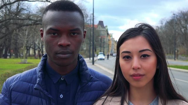 A young black man and a young Asian woman look at the camera in a street in an urban area - closeup on the faces