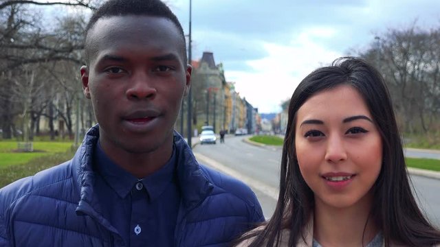A young black man and a young Asian woman talk to the camera in a street in an urban area - closeup on the faces