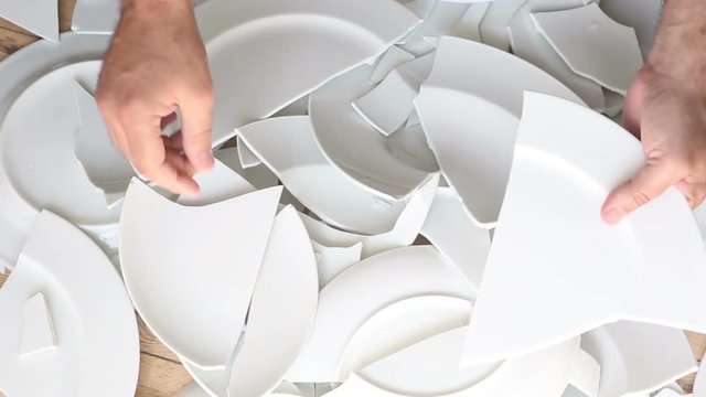 hands picking up broken white plates from wooden floor
