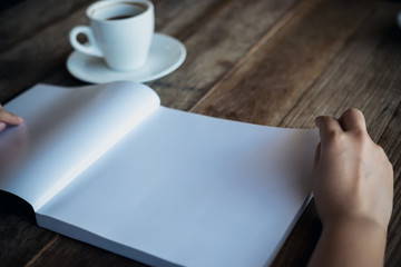 Closeup image of a woman reading and opening a white blank book with a white coffee cup on wooden table in vintage cafe