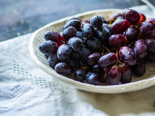 A bunch of ripe blue or purple grapes in a white bowl on a white linen napkin with a pattern on a dark background