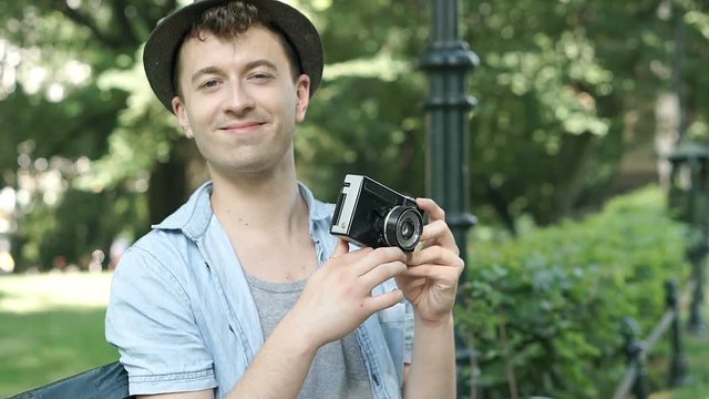 Young man in hat doing photos on old camera and sitting in the park, steadycam shot
