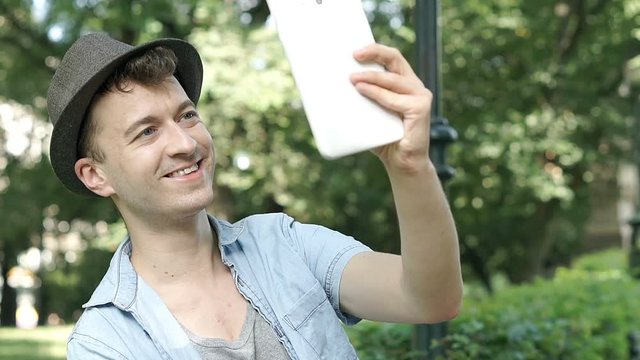 Young man in hat sitting in the park and doing selfies on tablet, steadycam shot

