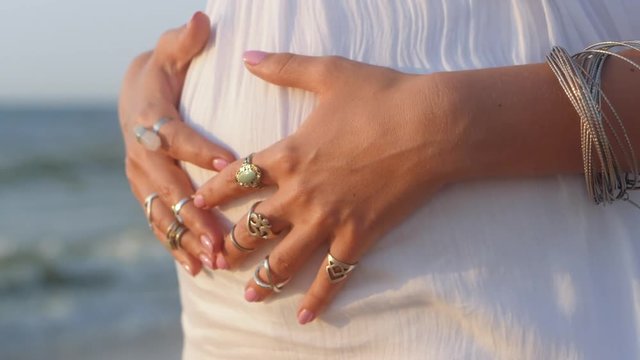 Pregnant young gypsy woman with Boho jewelry touching her belly. Girl in white long dress showing bohemian accessories near sea on the beach. Slow motion. Femininity concept.