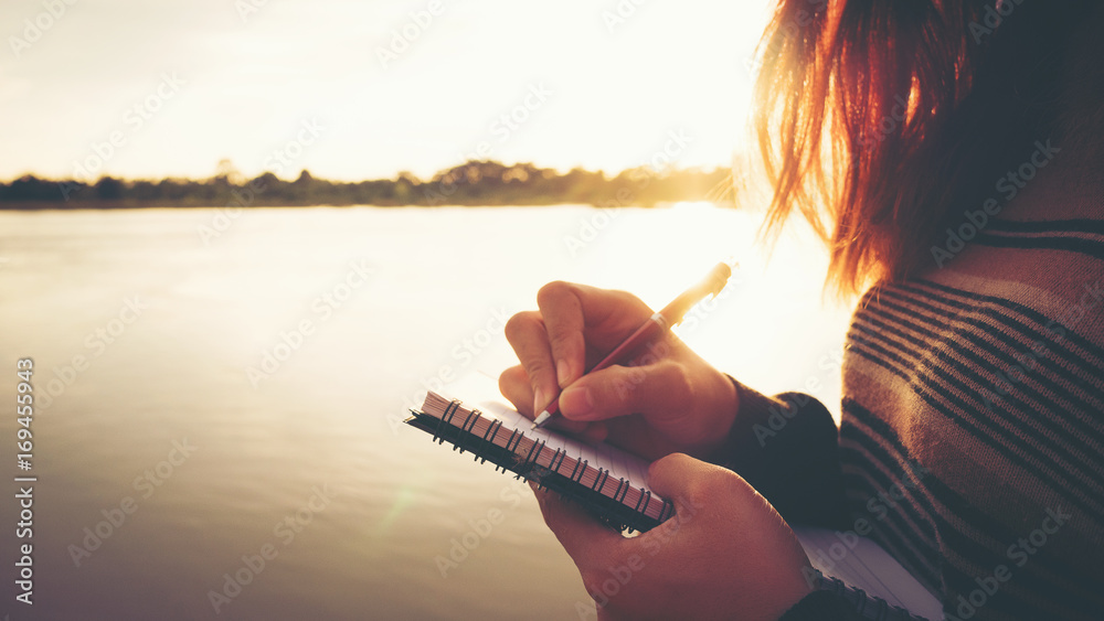 Wall mural close up hand of young woman with pen writing on notebook at riverside in the evening.