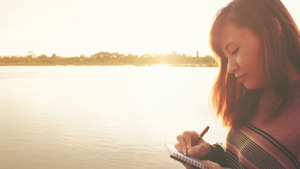 Fototapeta na wymiar Young woman with pen writing on notebook at riverside in the evening.