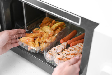 Woman putting container with fried potatoes, chicken meat and sausages into microwave oven in kitchen