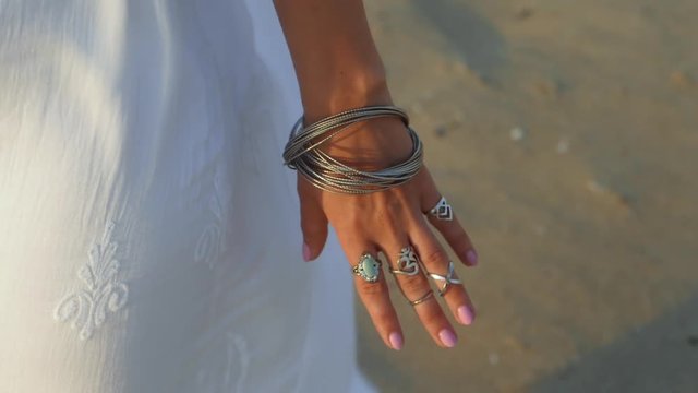 Beautiful young gypsy woman demonstrates boho jewelry. Girl in white long dress showing accessories near sea on the beach. Slow motion. Femininity concept.