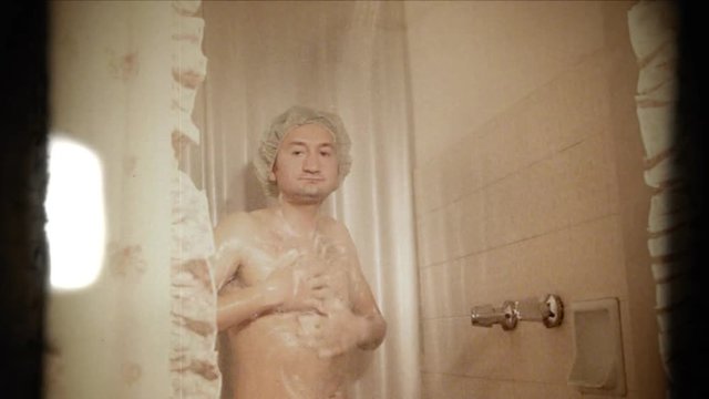 Fake 8mm amateur film: a funny slim ugly man singing in the shower, soaping himself while wearing a cap. Someone else is inside the tub, peeping at him. Gotcha! Close-up shot.
