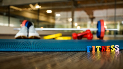 The word "Fitness" is built of colorful wooden letters with blurred back-up dumbbell, running shoes, weights and Mat.