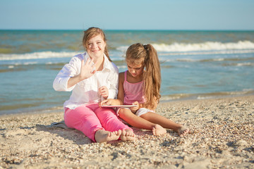 Girl seventeen-year-old  with Down syndrome and little girl on the beach play with the tablet