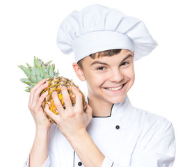Handsome teen boy wearing chef uniform holding ananas. Portrait of a happy cute male child cook with pineapple, isolated on white background. Food and cooking concept. 