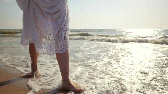 Legs of caucasian girl wearing white long dress and silver bracelets walking barefoot sand on sea beach, sun, lens, flare. Slow motion. Woman walks pass camera