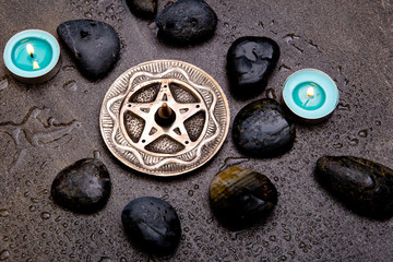 Incense burning in silver pentagram with blue candles and black rocks on gray slate background