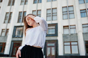 Portrait of a fabulous young successful woman in white blouse and broad black pants posing on the stairs with a huge white building on the background.