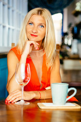 Young beautiful tanned blonde woman with blue eyes and res lips in ogange dress drinking wine from the wineglass while sitting near opened window in cafe somewhere in Mediterranean.