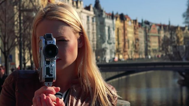 A young attractive woman shoots a video with a camera - closeup - a river, a bridge and a street of a quaint town in the blurry background