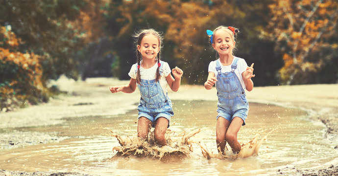 Happy funny sisters twins  child by girl jumping on puddles in rubber boots and laughing.