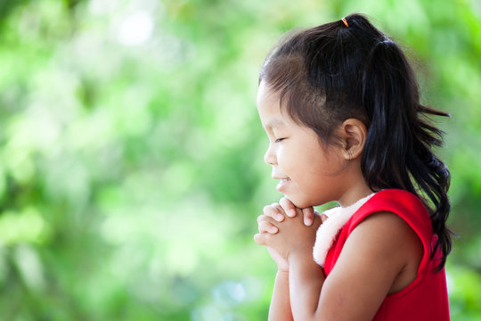 Cute asian little girl in christmas dress closed her eyes and folded her hand in prayer on green nature background. Faith and christmas concept.